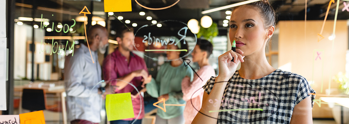 kobieta analizuje wykresy na szklanej ścianie, podczas gdy w tle toczy się dyskusja jej współpracowników
